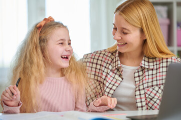 Happy child smiles at laptop screen while mother sits beside, positive learning moment. Supportive home study environment, engaging school preparation