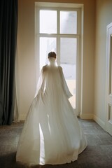 Fototapeta premium Bride in Elegant Wedding Dress Stands Before a Window Bathed in Natural Light