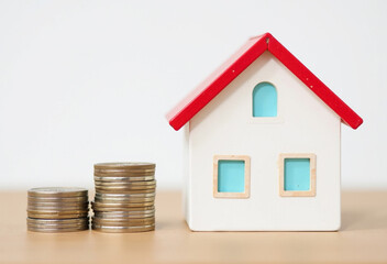 A small white house model with a red roof sits next to stacks of coins, representing homeownership and financial planning.
