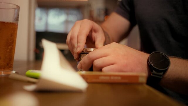 A man prepares cannabis for rolling by grinding dried buds with a metal grinder. The scene is set in a relaxed indoor environment with warm lighting, featuring rolling papers and a beverage.