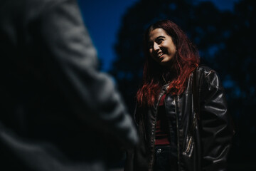 A young woman with red hair smiles while talking outdoors at night in soft lighting, embodying warmth and connection in an informal setting.