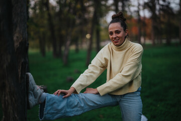 A cheerful young girl performs stretching exercises in a scenic green park setting. She is engaged...