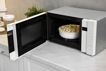 Open microwave oven with bowl of popcorn on white marble countertop in kitchen