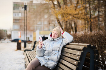 Fototapeta premium Portrait young Caucasian girl using smartphone, looking at mobile phone screen on winter walk in park