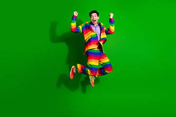 Young man jumps joyfully in a vibrant rainbow-striped suit on a bright green background, projecting happiness and colorful style.