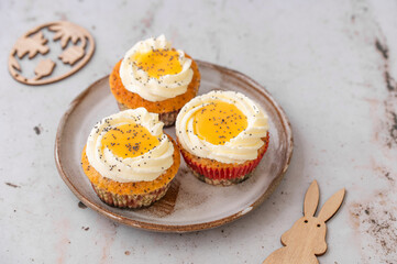 Lemon Poppy Seed Cupcakes with lemon curd and cream cheese topping. Cupcake in form of Fried Egg. Colourful Easter eggs, flowers on a stone background, top view. Easter holiday and festive food.