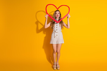 Young woman in a polka-dot outfit with a red heart-shaped frame on a vibrant yellow backdrop exuding joy and charm