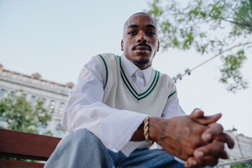 stylish Black man with tattoos and gold accessories sits on a bench, leaning forward confidently....