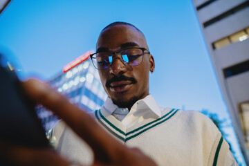 young Black man in glasses is deeply engaged with his smartphone against modern city skyline at...