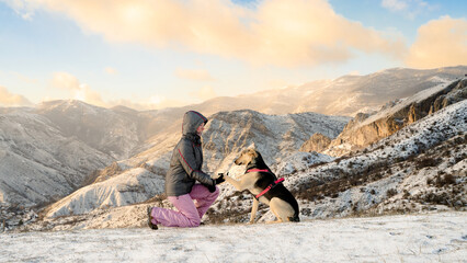 Fototapeta premium A dog giving his paw to his female owner against majestic winter mountains. A woman gives a command to her dog during a walk on a cold day. Good behavior, training with pet outdoors. Bonding and fun.