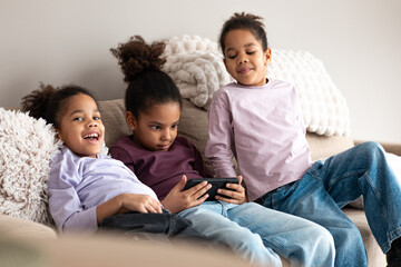 Fototapeta premium Three happy sisters lounging on a couch, engaged with a smartphone