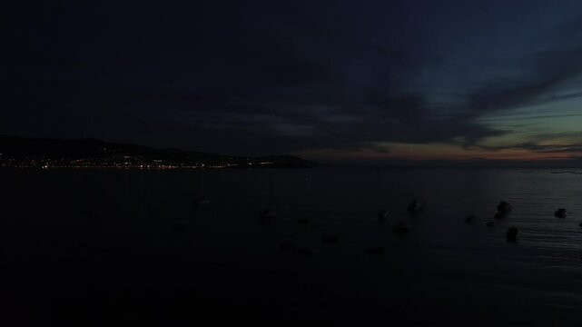 Aerial view descending over Panx&oacute;n beach after blue hour. With its anchored boats, the illuminated Panx&oacute;n harbor on the right, and Baiona&rsquo;s lights in the distance. Calm sea reflects harbor lights.