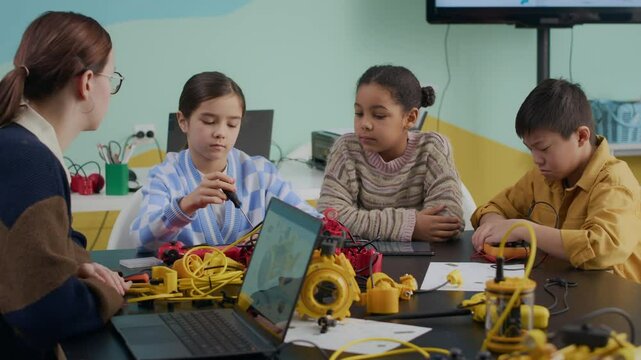 Medium shot of young Caucasian female teacher in casualwear sitting at table with Gen Alpha multiethnic kids and explaining how to construct robot using digital drawing on laptop screen