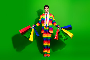 Cheerful young man in a vibrant rainbow-patterned suit holding colorful shopping bags against a green background