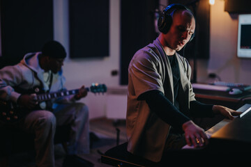 Two focused musicians, one playing piano and one on guitar, recording in a dimly lit studio. Both wear headphones, immersed in creating music. The ambiance is creative and concentrated.