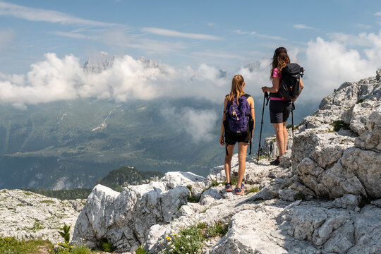 Two women with backpacks hike on a rocky mountain ridge, looking out at a valley and distant, cloud-covered peaks.Flower path, Julian natural Park, Gilberti hut, Sella Nevea, Canin mount.