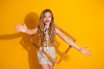 Excited young woman in fashionable outfit framed by a wooden square on a vibrant yellow background sharing positivity with open arms