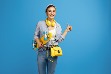 Naklejka premium Modern young woman hold skateboard over blue background. Woman showing thumb up