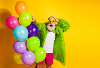 Energetic man in a green fur coat with balloons against a vibrant yellow background, expressing joy and highlighting colorful fashion
