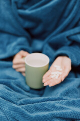 Woman wrapped in blue blanket holding cup of tea and pills in hand