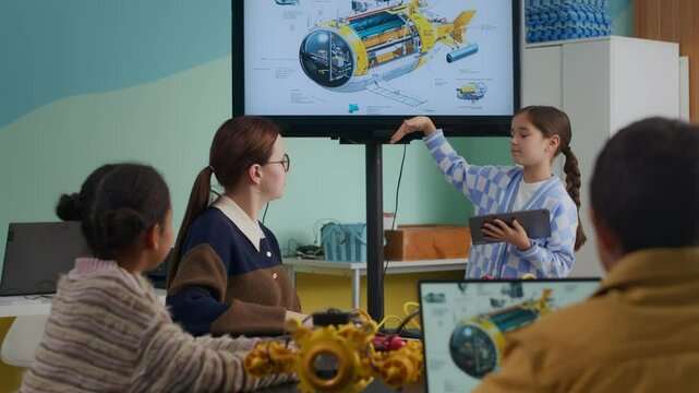 Asian girl holding digital tablet explaining structure of robot to young Caucasian female teacher and children sitting at table during STEM education lesson in modern well-equipped classroom