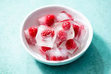 Frozen ice cubes with fresh raspberries in a white bowl. Blue background. Close up.