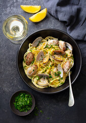 Spaghetti alle vongole, seafood pasta with clams in a black bowl and glass of white wine. Dark background. Top view.