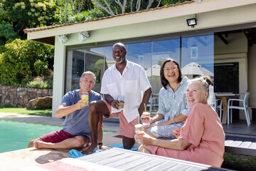 Diverse senior friends enjoying drinks by pool, laughing and relaxing in sunny backyard