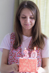 A young 30-year-old woman celebrates her 30th birthday with a cake with candles 30. Natural face portrait of a young caucasian woman, no retouching skin. Brunette girl, naturalness.