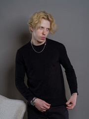 A young man with white wavy hair, wearing a black sweater, seated in a chair against a gray background. The simple yet stylish composition highlights his relaxed and confident demeanor.
