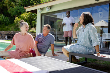 Senior friends relaxing by pool, enjoying conversation and drinks outdoors