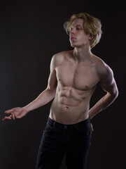Artistic portrait of a young man with wavy blonde hair against a dark background. Bare shoulders, hand on chest. Thoughtful gaze, melancholic expression. Soft lighting highlights facial features
