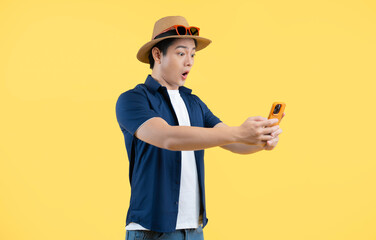 Portrait of young Asian man wearing tourist clothes and using phone on yellow background