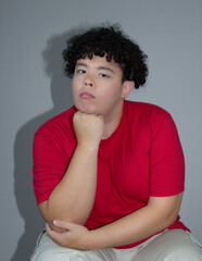 Young man with curly hair wearing a red t-shirt, resting his chin on hand while looking at camera....