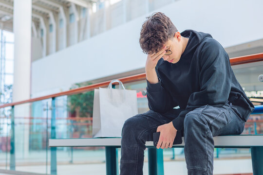 eenager in black hoodie sitting on a bench with head in hands, feeling stressed or overwhelmed, sensory overload, autism, ADHD, bullying, mental health in a bright shopping mall. Copy space