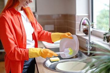 Woman with orange hair cleaning dishes in a bright kitchen during the day while wearing rubber gloves and a cheerful smile