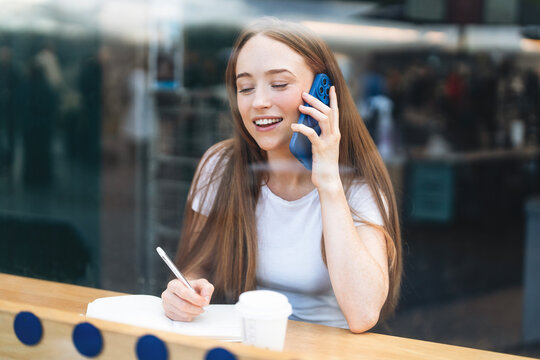 Young woman engaged in a thoughtful conversation on her phone while taking notes at a cafe during the afternoon hours in a bustling urban environment