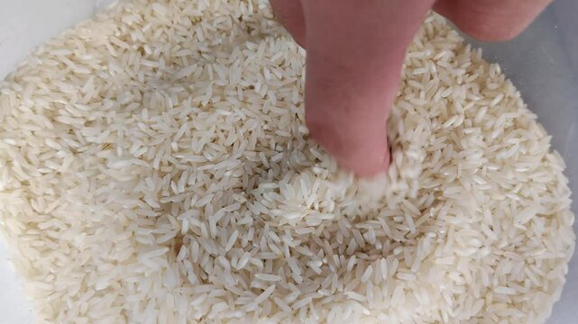 A hand that is immersed and appears to be moving its fingers over dry, uncooked grains of white rice.