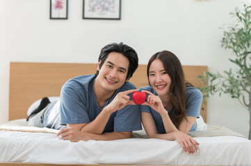 Young Asian couple in love laying on bed and holding red heart enjoying spending time together, Celebrating Valentine's Day