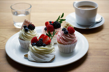 Cupcakes and mini pavlova cakes decorated with fresh berries and white chocolate on a large white plate, a cup of coffee and a glass of water on wooden table. Selective focus.