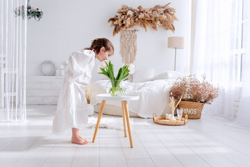 Young girl in a white dress is bending over to smell a bouquet of fresh white tulips placed on a small table in a bright, bohemian-style bedroom with a comfortable bed and natural decorations