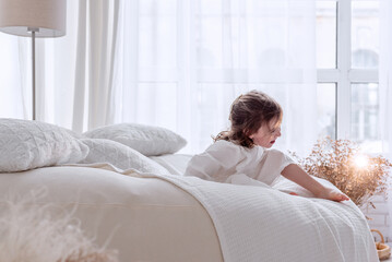 Cute little girl wearing a white dress joyfully playing on a bed in a bright white bedroom, embracing the cheerful atmosphere of a playful morning at home