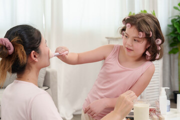 Fototapeta premium Caucasian girl playfully applying makeup on Asian mother’s face with a brush. Warm and happy family bonding moment, learning beauty techniques together in a cozy home setting.