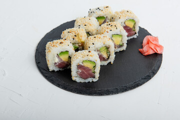 Close-up of sushi California rolls filled with tuna, avocado, and sesame seeds served on a black slate plate, accompanied by pickled ginger on isolated white background