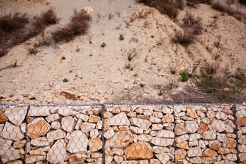 Close-up of a gabion wall made of irregular stones held by a metal mesh, set against a rugged hillside with sparse vegetation and trees in the background.