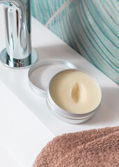 Opened body butter jar with a lid on white basin near brown towel and faucet close up, cosmetic mockup