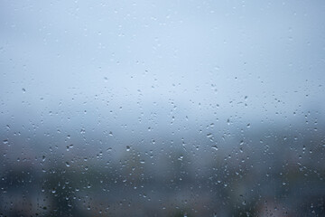 Raindrops on glass window with blurred cityscape in the background. Wet transparent surface covered with water droplets on a rainy day. Abstract weather and nature concept for design and print