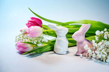 Two Easter bunnies from ceramic with a bouquet of pink tulips against a light blue background, holiday greeting card, copy space, selected focus