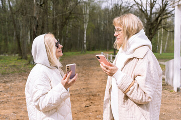 Two senior women standing in a park and talking while holding their smartphones. Dressed in stylish neutral-tone outfits with hoodies and sunglasses, they represent active aging and communication.