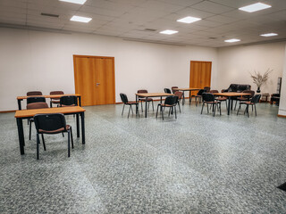 A spacious conference room with wooden tables and black chairs arranged for meetings or events. The minimalist interior features plain walls, wooden doors, and overhead lighting.